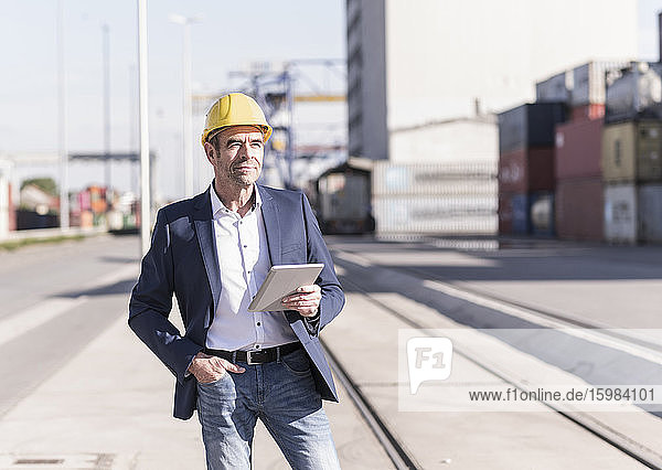 Portrait of content businessman wearing safety helmet with digital tablet at industrial site