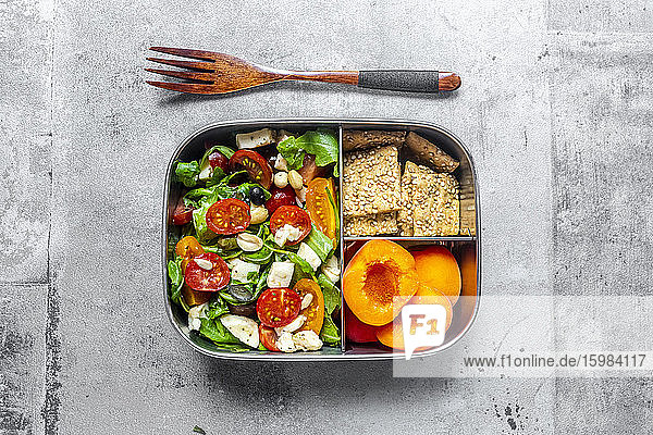 Lunch box with rocket salad with colored tomatoes  mozzarella and nuts  crispbread and apricots  wooden fork on concrete surface