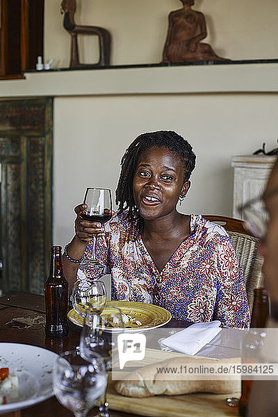 Lächelnde junge Frau mit Weinglas in der Hand  die während des Mittagessens am Esstisch sitzt