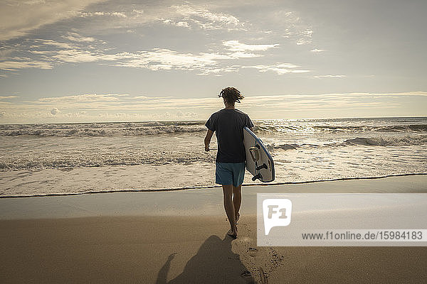 Costa Rica  Provinz Puntarenas  Puntarenas  Rückansicht eines männlichen Surfers  der mit einem Surfbrett in der Hand auf das Meer zugeht
