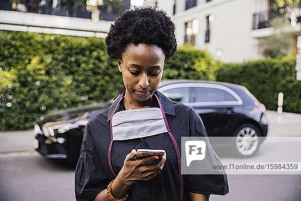 Portrait of young woman with protective mask standing on the street looking at cell phone