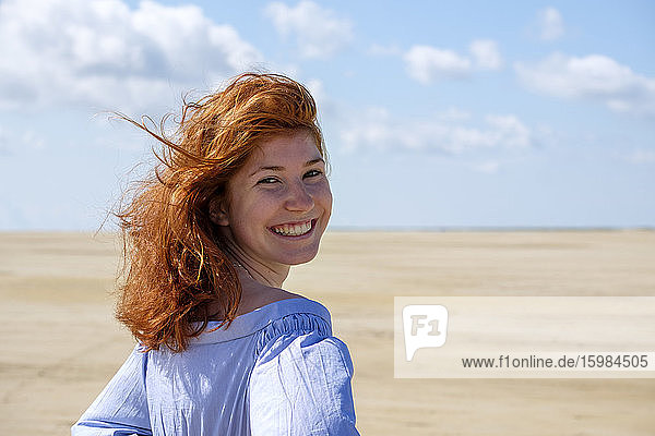Lächelndes Teenager-Mädchen  das über die Schulter schaut  während es am Strand gegen den Himmel an einem sonnigen Tag steht