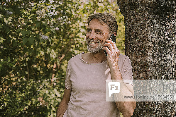 Smiling mature man talking on mobile phone while leaning on tree at garden