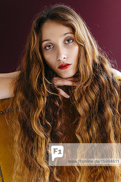 Porträt einer jungen Frau mit langen braunen gewellten Haaren  die sich auf einen Stuhl vor einem farbigen Hintergrund lehnt