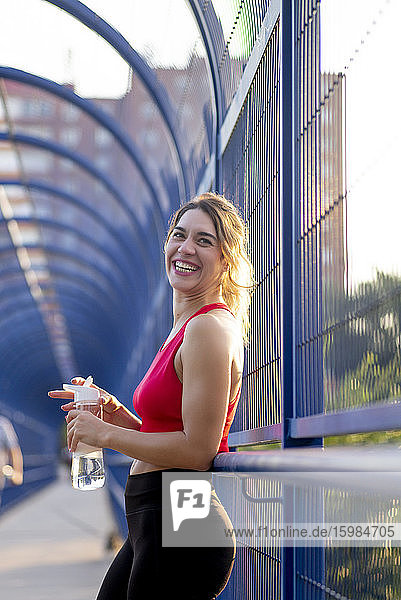 Lachende Sportlerin  die eine Wasserflasche hält und wegschaut  während sie auf einer überdachten Brücke in der Stadt steht
