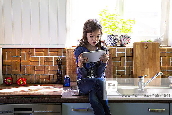 Girl sitting on sink in the kitchen looking at digital tablet