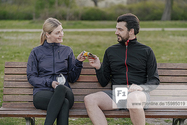 Lächelnde Frau  die einem Mann eine Gesichtsmaske gibt  während er während der Quarantäne im Park auf einer Bank sitzt