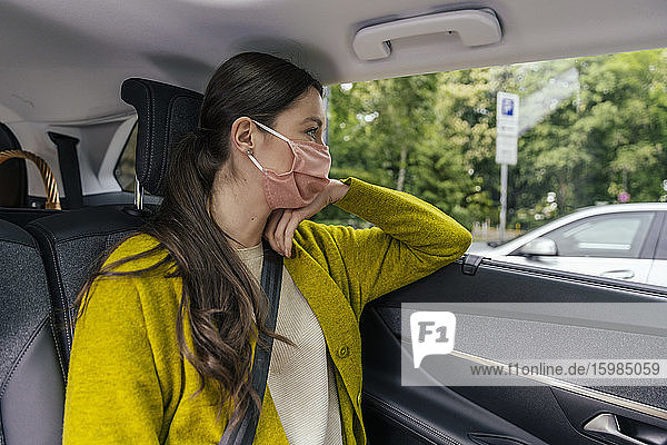 Junge Frau mit Schutzmaske sitzt auf dem Rücksitz eines Autos und schaut aus dem Autofenster