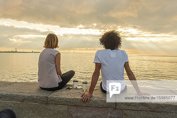 Rückansicht eines Paares  das auf einer Stützmauer am Meer sitzt  gegen einen bewölkten Himmel bei Sonnenuntergang