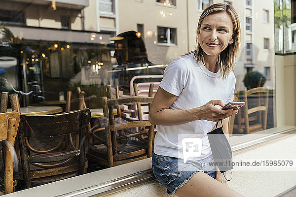 Porträt einer Frau mit Smartphone und Schutzmaske in der Hand  die vor einem Café wartet