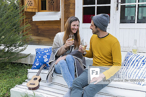 Glückliches Paar mit Weingläsern in der Hand  das sich im Sitzen an einer Blockhütte unterhält