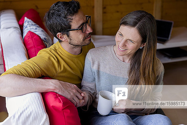 Mann betrachtet nachdenkliche Freundin mit Kaffee in der Hand  während er auf dem Sofa in einer Blockhütte sitzt
