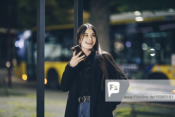 Glückliche junge Frau  die wegschaut  während sie mit ihrem Smartphone nachts im Freien steht