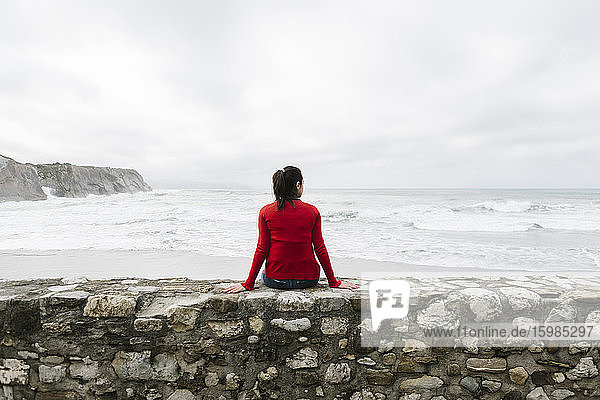 Rückansicht einer entspannten Touristin  die auf einer Stützmauer sitzt und das Meer gegen den Himmel betrachtet  Itzurun  Zumaia  spanisches Baskenland  Spanien