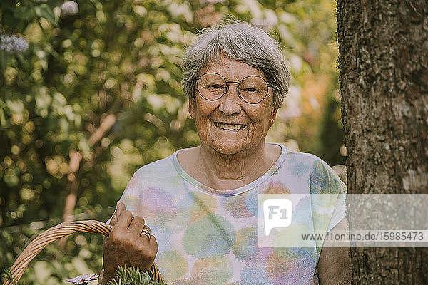 Happy retired senior woman holding basket while standing by tree at garden