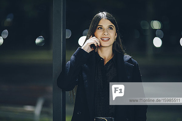 Smiling beautiful young woman looking away while talking on mobile phone outdoors at night