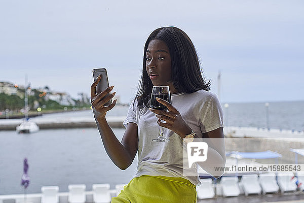 Porträt einer erstaunten jungen Frau auf einer Terrasse mit einem Glas Rotwein  die auf ihr Mobiltelefon schaut