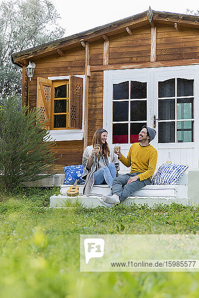 Glückliches Paar mit Weingläsern in der Hand  das sich auf den Stufen einer Blockhütte unterhält