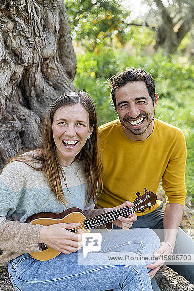 Fröhliche Frau spielt Gitarre  während sie mit ihrem Freund an einem Baum in der Natur sitzt