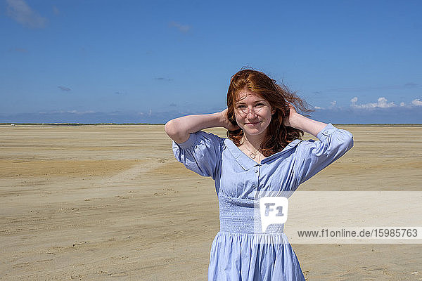 Portrait of smiling teenage girl wearing blue dress standing at beach against sky on sunny day