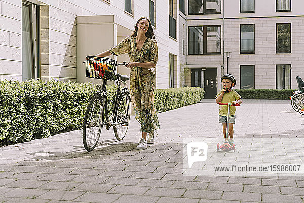 Smiling mother with bicycle and son with scooter in the city