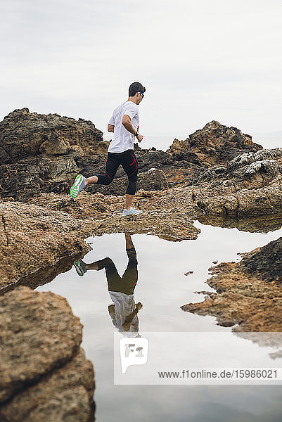 Full length of male trail runner on rocks while running by his reflection over water at coast  Ferrol  Galicia  Spain