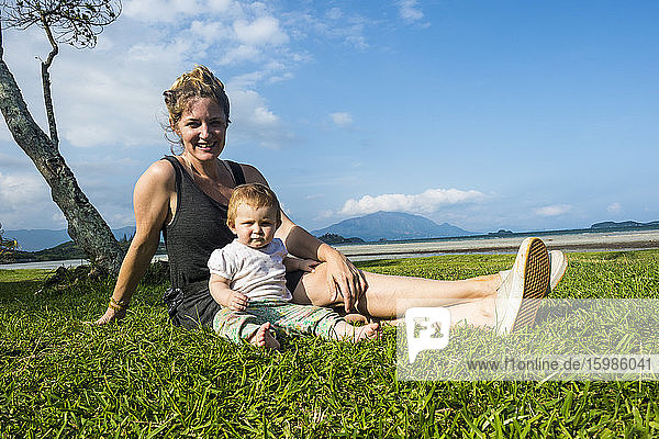 Porträt einer lächelnden Mutter mit ihrer süßen kleinen Tochter auf einer Wiese vor blauem Himmel  Noumea  Neukaledonien
