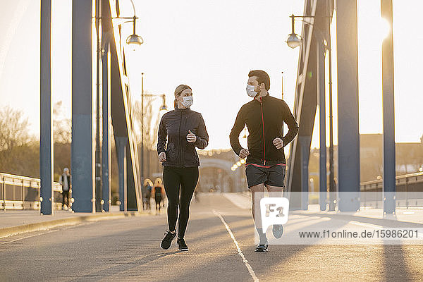 Junges Paar mit Gesichtsmasken läuft bei Sonnenuntergang auf einer Brücke in der Stadt