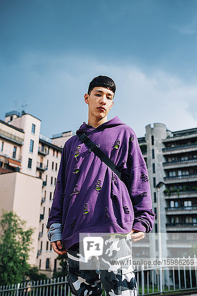 Serious young man standing against blue sky in city