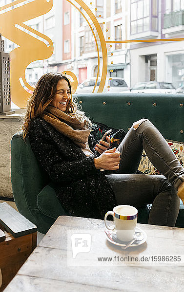 Lachende Frau mit Smartphone auf der Couch in einem Café sitzend