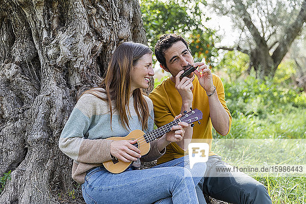 Happy couple playing musical instruments while sitting by tree trunk