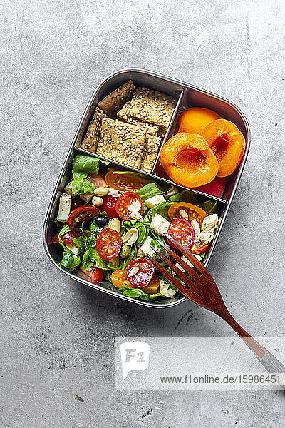Lunchbox mit Rucolasalat mit bunten Tomaten  Mozzarella und Nüssen  Knäckebrot und Aprikosen  Holzgabel auf Betonfläche