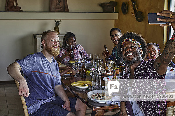 Lächelnder Mann  der ein Selfie mit Freunden macht  die während des Mittagessens am Esstisch sitzen