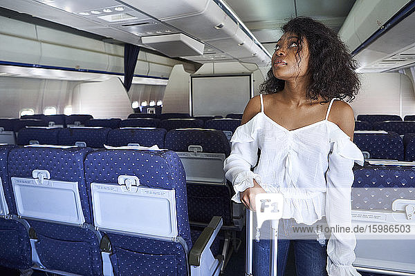 Young woman searching seat while standing with luggage in airplane