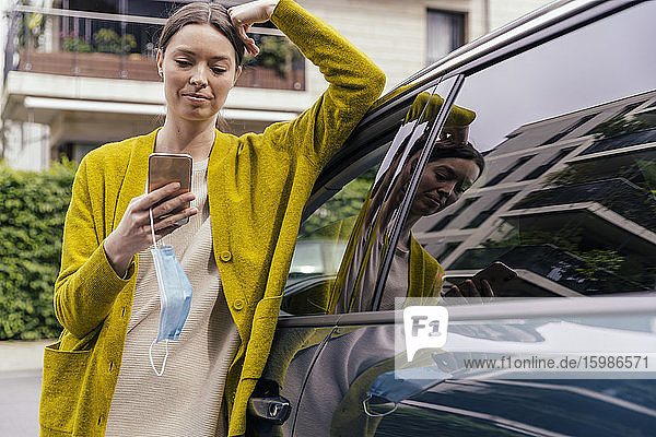 Porträt einer jungen Frau mit Schutzmaske  die sich an ihr Auto lehnt und auf ihr Mobiltelefon schaut