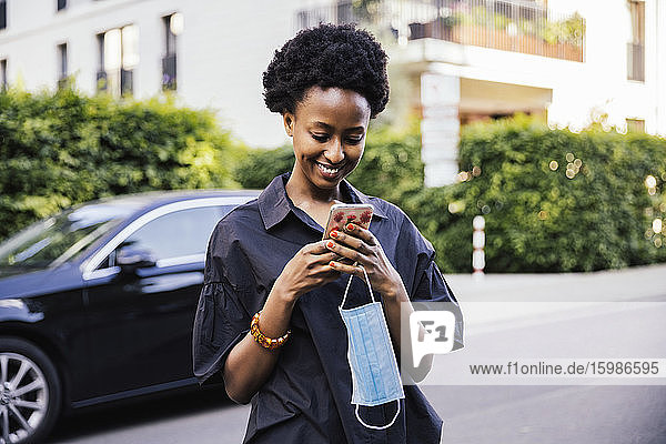 Porträt einer lächelnden jungen Frau mit Schutzmaske  die auf der Straße steht und auf ihr Mobiltelefon schaut