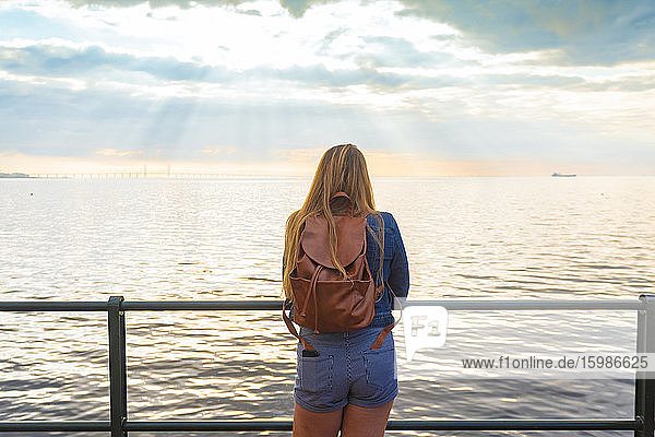 Rear view of woman with backpack standing at railing by sea against sky