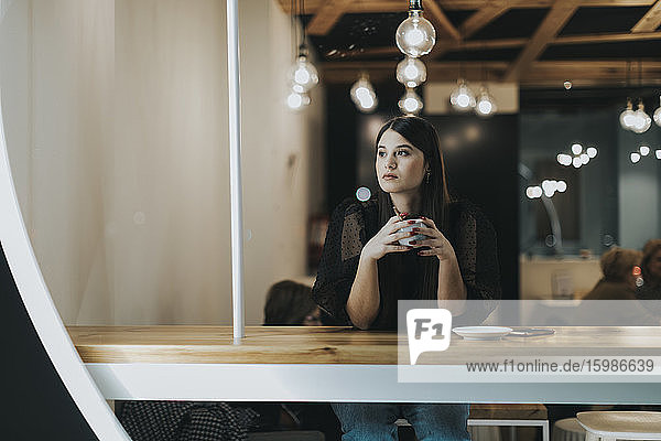 Schöne junge Frau schaut weg  während sie mit einer Kaffeetasse im Café sitzt und nachdenkt
