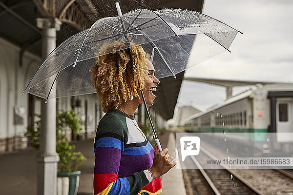 Seitenansicht einer fröhlichen jungen Frau  die mit einem Regenschirm am Bahnhof steht und sich über den Monsun freut