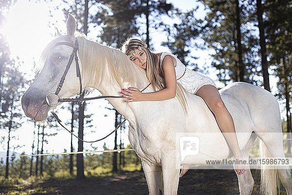 Full length portrait of young woman leaning on white horse in forest