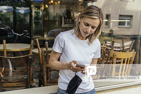 Porträt einer Frau mit Schutzmaske in der Hand  die vor einem geschlossenen Café Textnachrichten verschickt