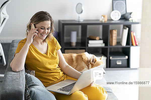 Frau mit Hund benutzt Laptop und Mobiltelefon im Wohnzimmer zu Hause