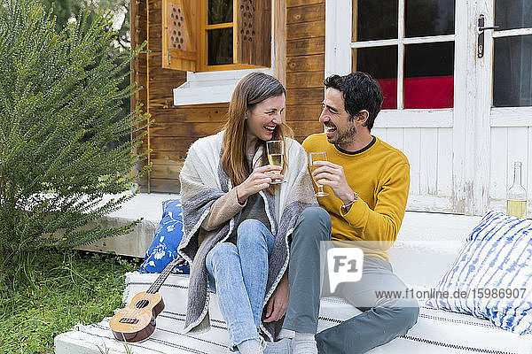 Laughing couple holding wineglasses while sitting against log cabin
