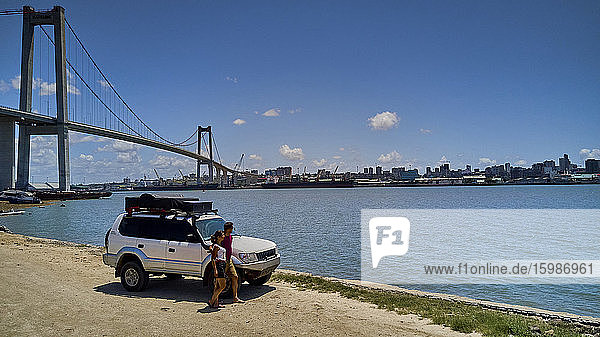 Mosambik  Katembe  Erwachsenes Paar steht bei 4x4 Auto und bewundert die Aussicht auf die Maputo Bucht mit der Stadt und der Maputo-Katembe Brücke im Hintergrund