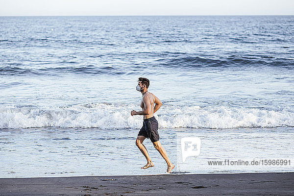 Shirtless Mitte erwachsenen Mann trägt Gesichtsmaske beim Laufen am Strand bei Sonnenuntergang
