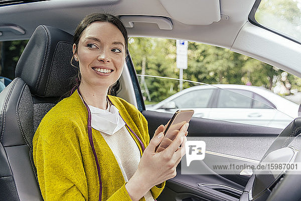 Porträt einer lächelnden Frau im Auto mit Schutzmaske und Smartphone
