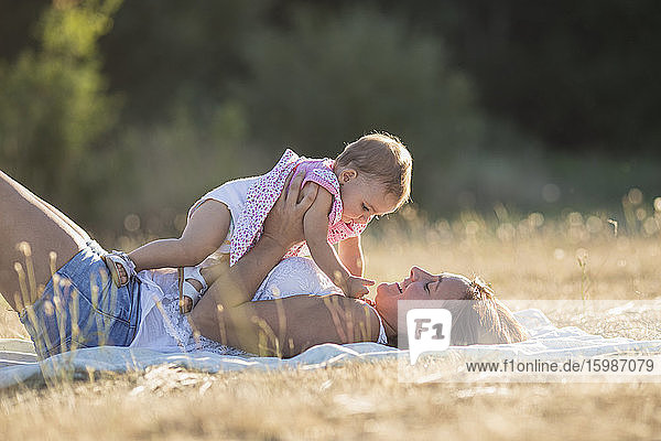 Glückliche Frau  die ein kleines Mädchen trägt  während sie auf einer Decke auf einer Wiese in der Sonne liegt