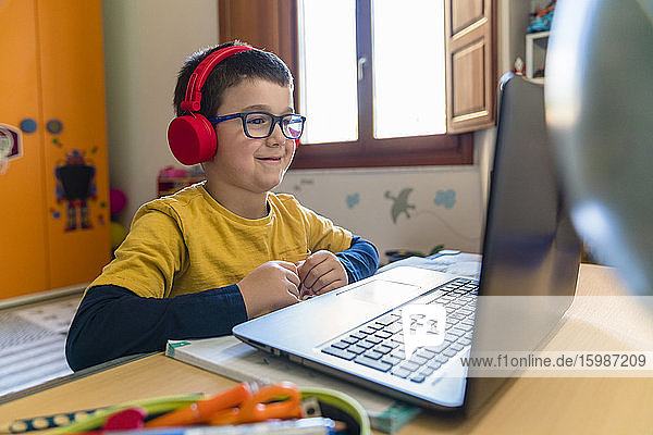 Smiling male student listening through headphones while looking at laptop during homeschooling