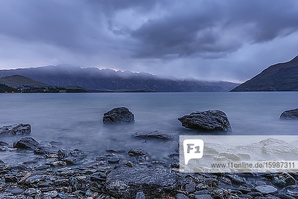 New Zealand  Otago  Cloudy sky over rocky shore of Lake Wakatipu at dawn with Remarkables in background