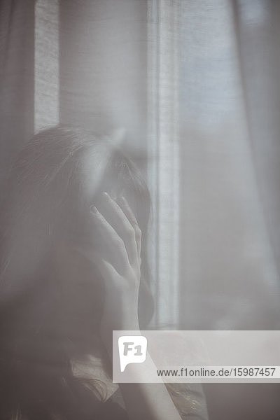 Junge Frau versteckt sich hinter weißem Fenstervorhang